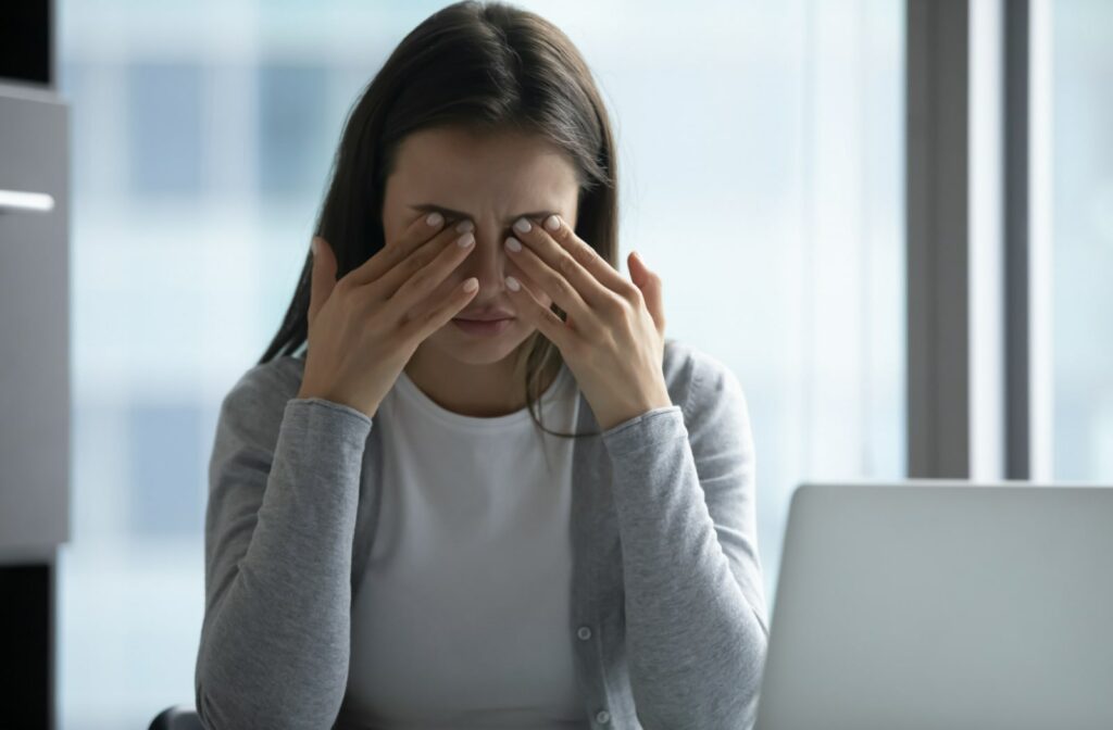 A young woman sitting in front of her laptop computer is massaging her eyes to relieve the blurriness of her sight due to dry eyes