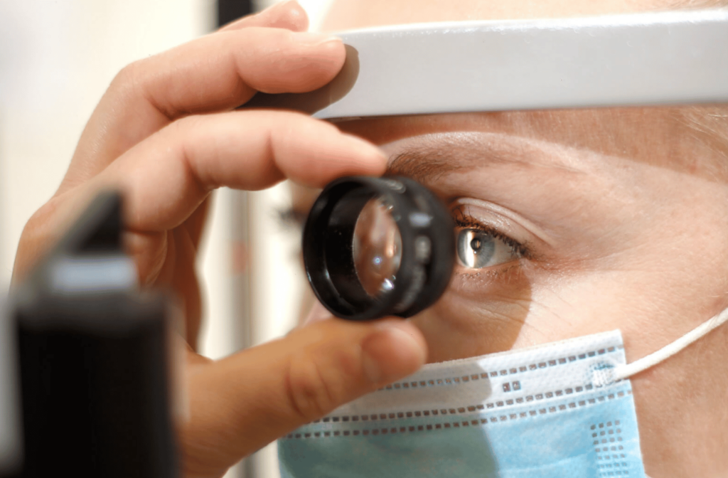 Close-up view of a woman's face, wearing a medical face mask, as an optometrist examines her left eye using a small magnifying glass.