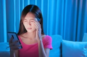 A woman sitting in a dark room squints due to eye strain from looking at her phone screen too long.