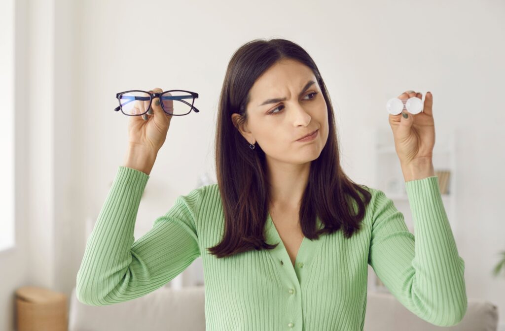 Woman with questionable expression looks at contact lenses she holds in one hand and has glasses in other hand.