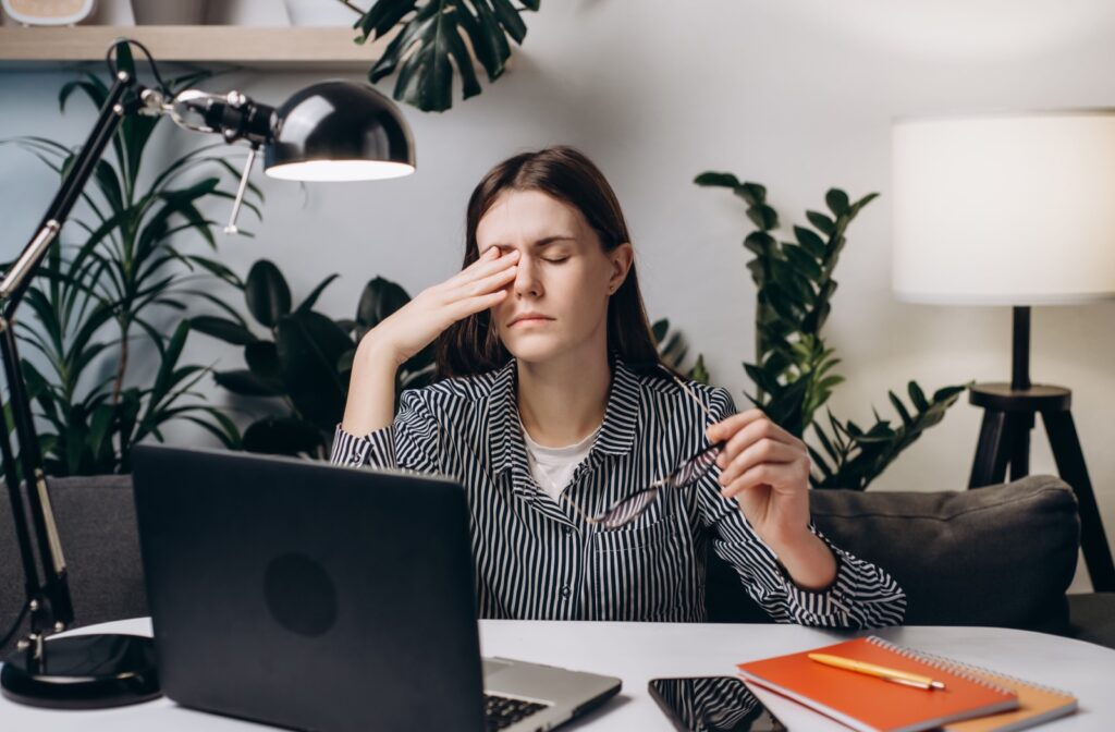 A young woman sits in her home office working on a laptop. She holds her glasses and massages her nose to reduce eye strain.