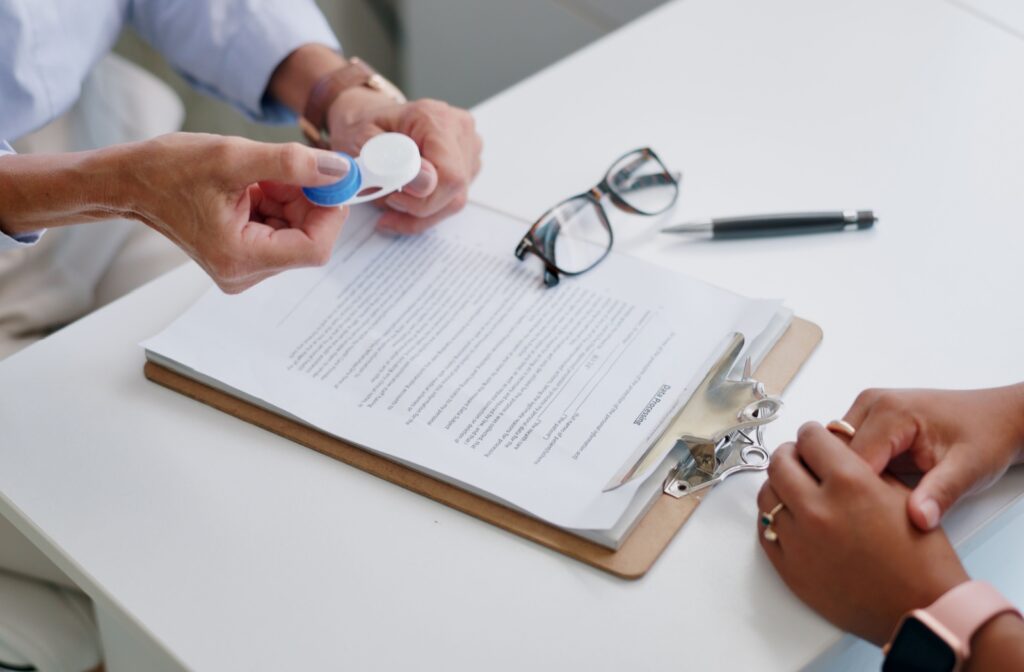 An optometrist holds out a case of contact lenses while explaining the importance of routine exams to a patient during a consultation. In the background, there are glasses and paperwork on the desk.