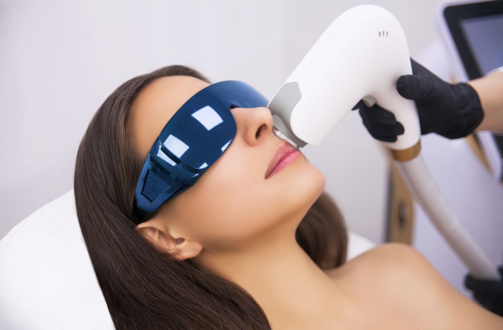 Woman receiving an IPL facial treatment while wearing protective goggles, with a technician using a handheld light-based device on her upper lip.